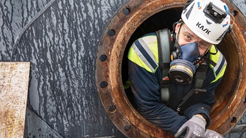 Man med gasmask tittar ut från arbete i slutet och trångt utrymme.