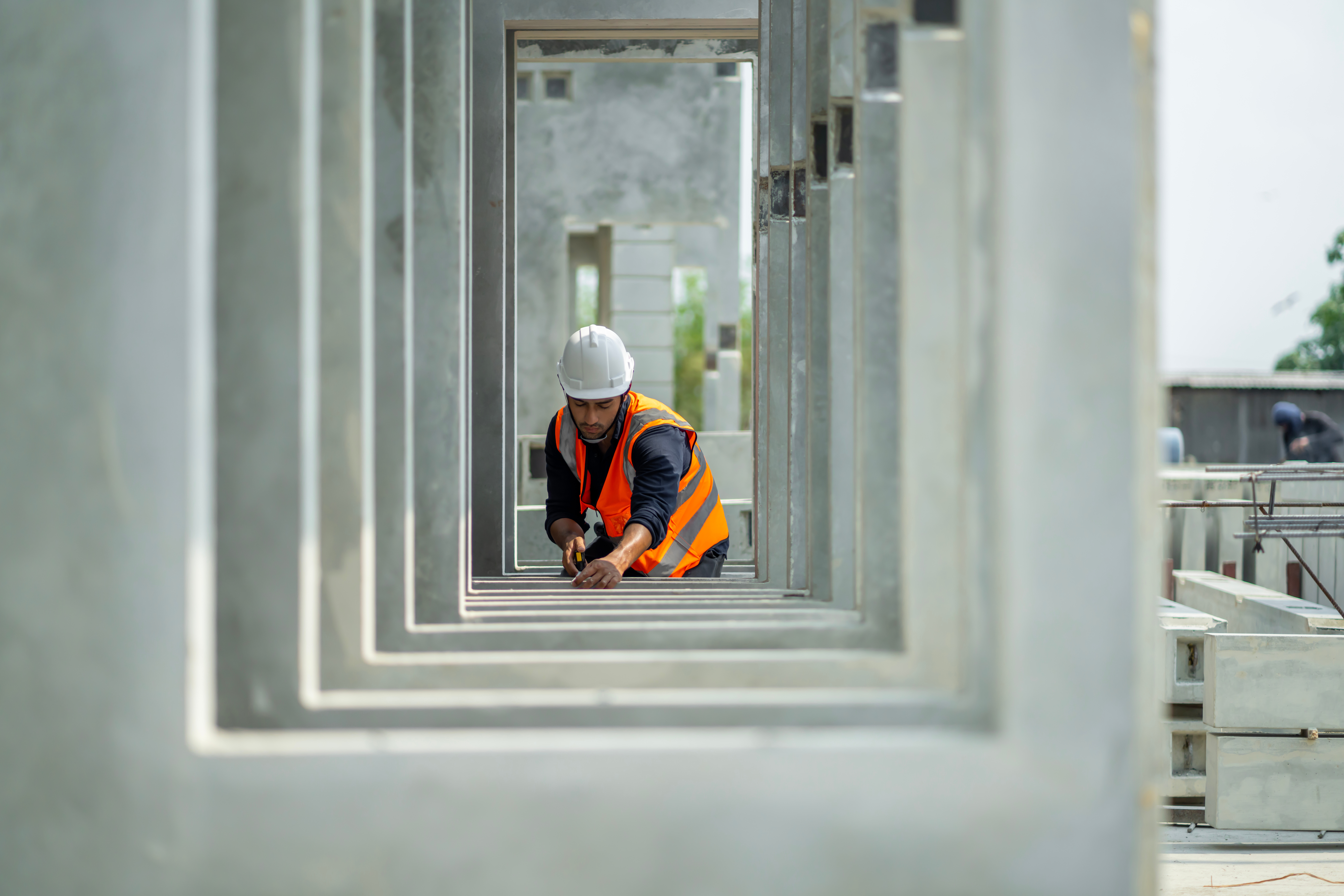 A construction worker in an orange safety vest and white helmet kneels inside a concrete structure, focusing on a task.