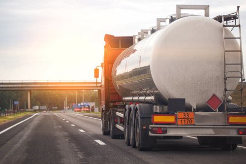 Tanker truck transporting liquid cargo on a highway during sunset, with a bridge in the background