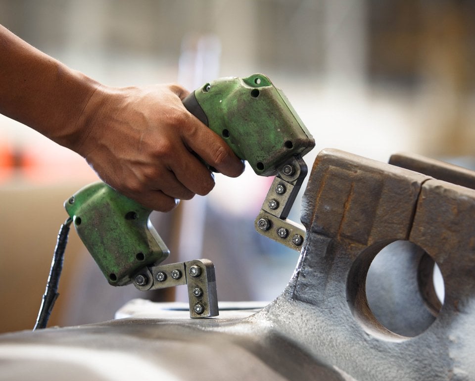 Hand som håller ett grönt elektriskt verktyg mot en metallkomponent för att magnetisera provobjektet, en metod inom oförstörande provning som kallas magnetpulverprovning och görs för att upptäcka sprickor i stål och järn.