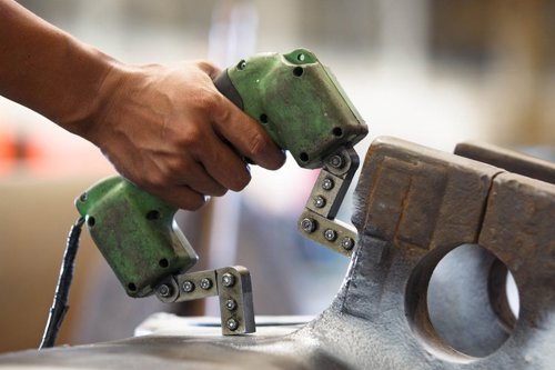 Hand som håller ett grönt elektriskt verktyg mot en metallkomponent för att magnetisera provobjektet, en metod inom oförstörande provning som kallas magnetpulverprovning och görs för att upptäcka sprickor i stål och järn.