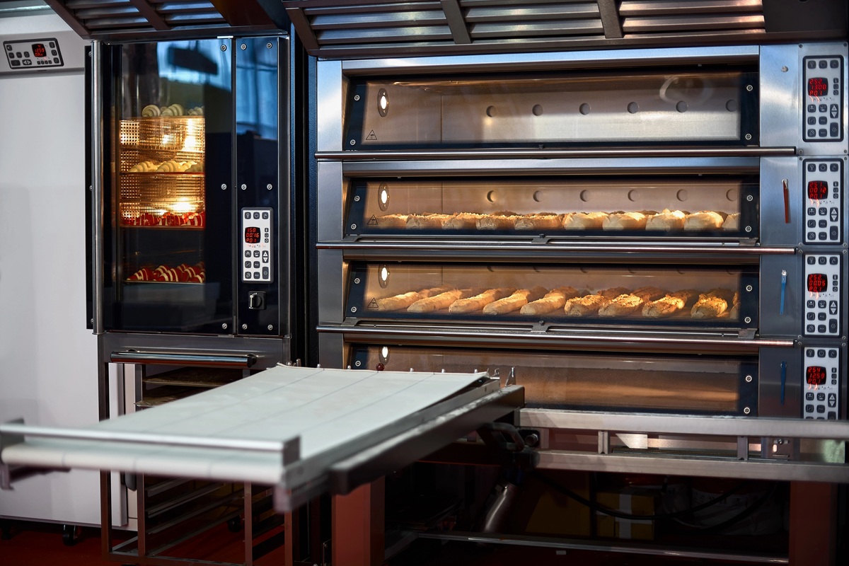 Commercial bakery oven with multiple trays of bread rolls baking inside, alongside a separate heating unit and a metal worktable.