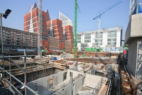Bouwplaats met graafwerkzaamheden, omringd door moderne gebouwen en hijskranen in een stedelijke omgeving.