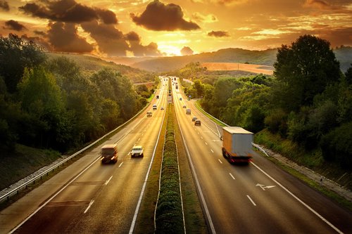 Snelweg met meerdere rijstroken omgeven door groene bomen en heuvels bij zonsondergang.