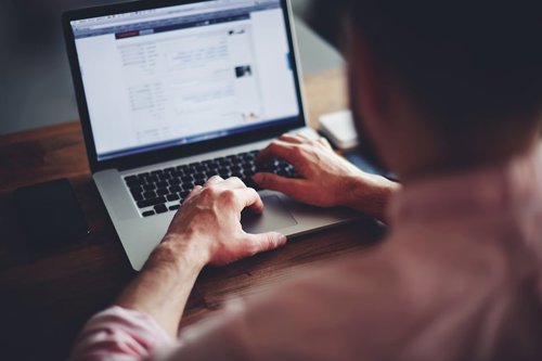 Man die typt op een laptop met een smartphone naast hem.