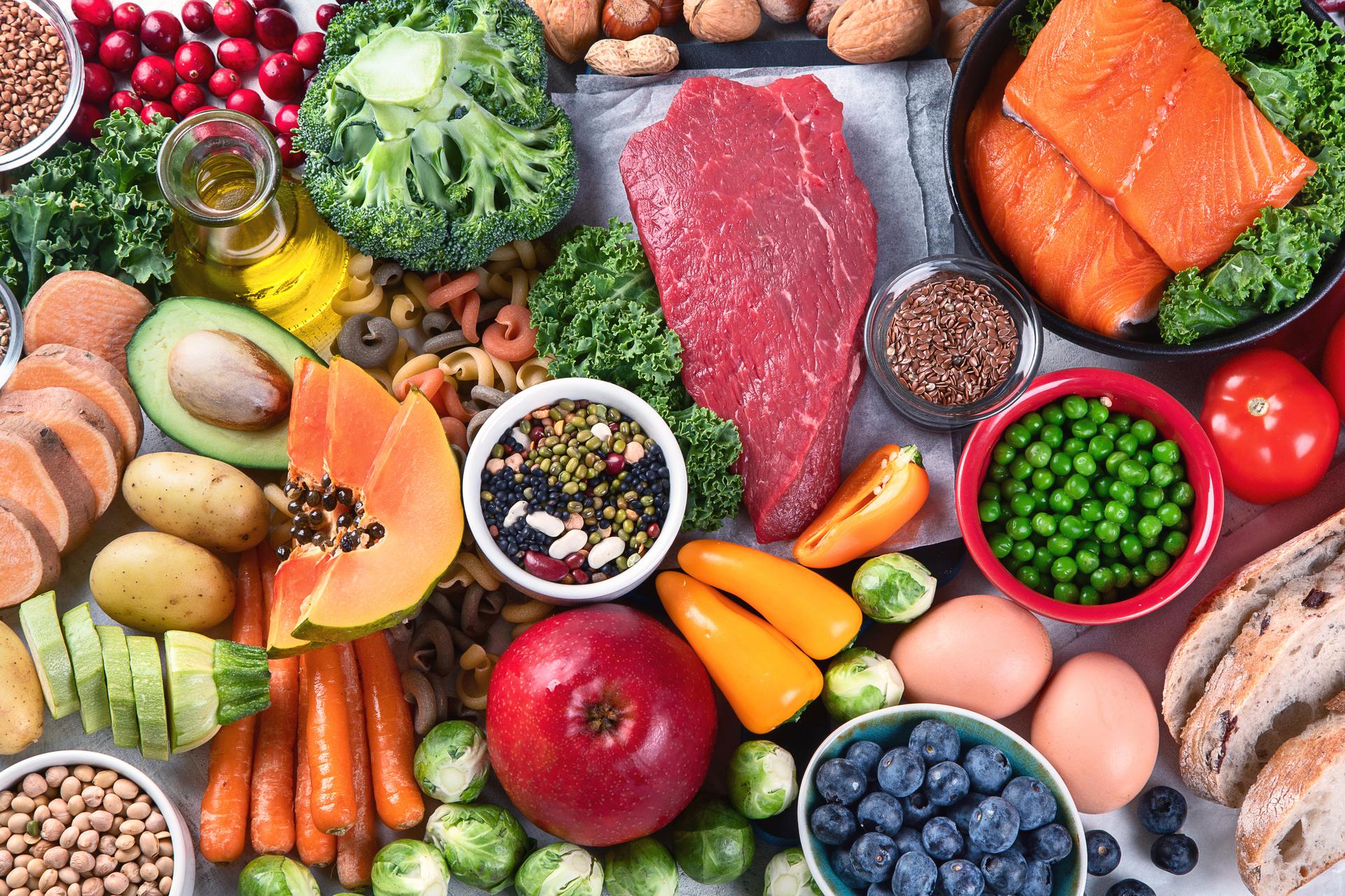 Assortment of fresh healthy foods including salmon, beef, eggs, vegetables, fruit, beans, nuts, and bread on a table.