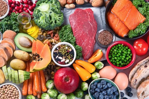 Assortment of fresh healthy foods including salmon, beef, eggs, vegetables, fruit, beans, nuts, and bread on a table.