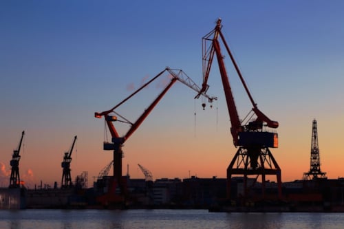 Hamn med stora industriella kranar mot en solnedgångshimmel vid vattnet.