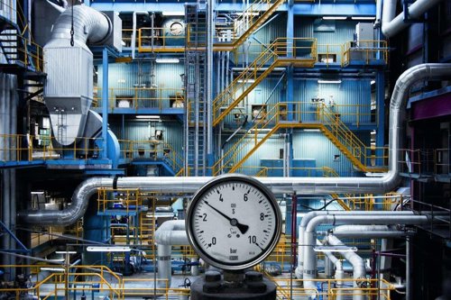 Tryckmätare i förgrunden av en industriell anläggning med rör och metallstrukturer - Kiwa erbjuder en kurs i avsäkring och dess processdesign.
