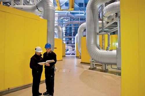Två personer i skyddshjälm inspekterar ett industriutrymme med stora ventilationsrör och gula väggar - Kiwa erbjuder en kurs till dig som har fortlöpande tillsyn av trycksatta anordningar där utbildningen varvar teori med praktik.