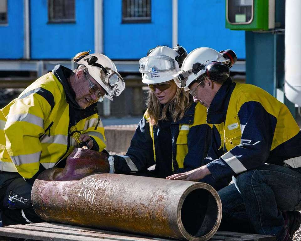Tre personer från Kiwa i skyddsutrustning inspekterar ett stort metallrör utomhus, med en blå byggnad i bakgrunden.