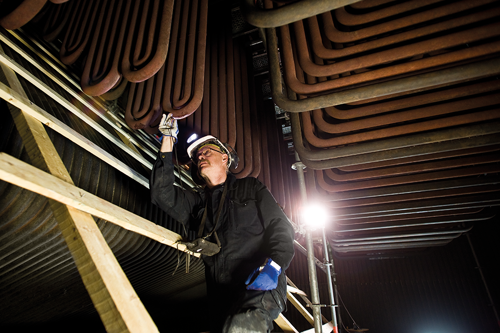 Inspektör i skyddsutrustning kontrollerar metallrör i en industriell anläggning, med byggnadsställningar och belysning i närheten.