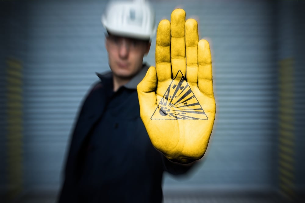 A person wearing a black shirt and a white safety helmet holds up a yellow-gloved hand with an intricate geometric design on the palm, standing in front of a blurred gray background.