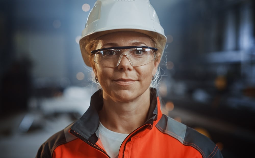 Portret van een vrouw in een witte veiligheidshelm, veiligheidsbril en oranje werkjas in een industriële omgeving.