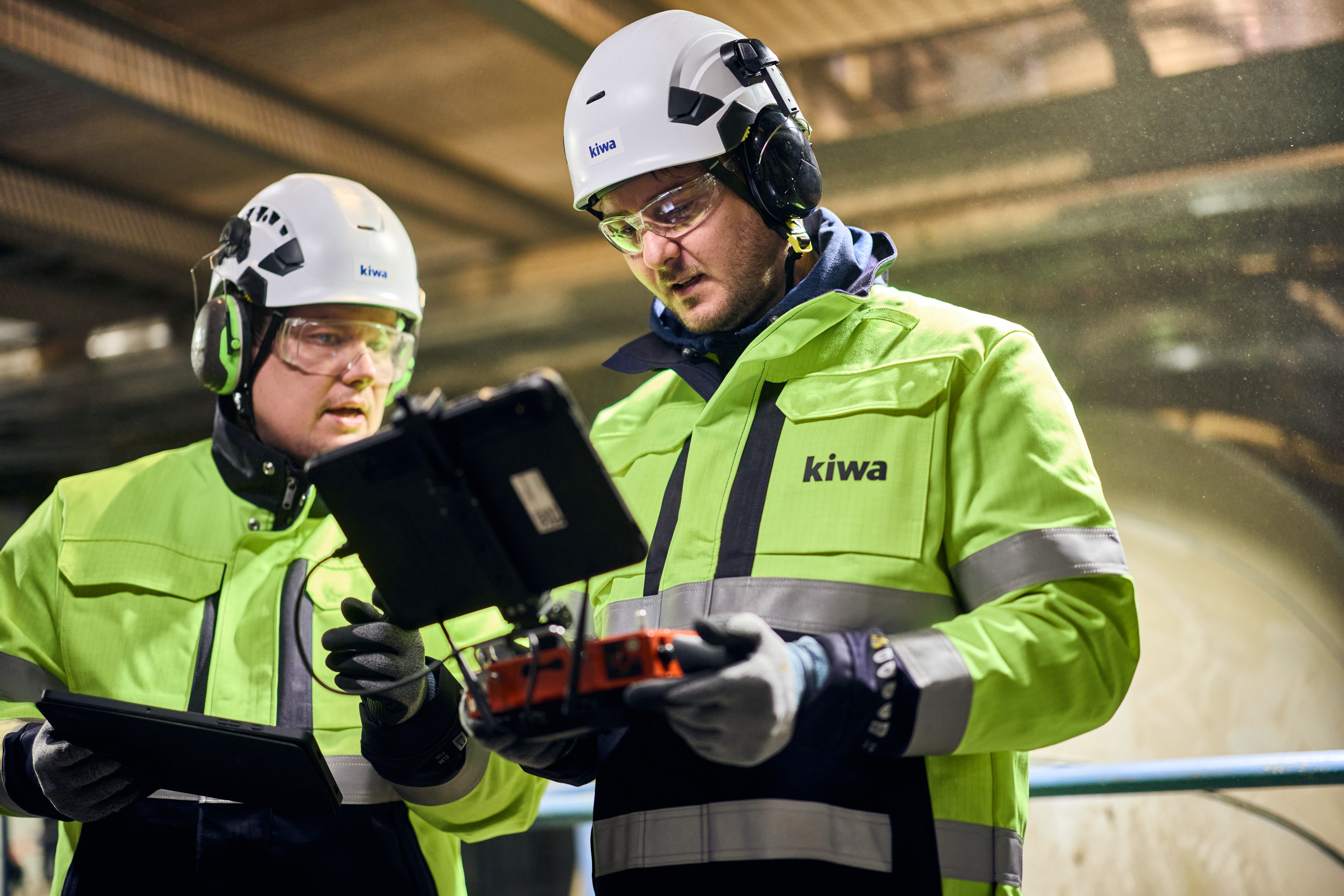 Two workers in high-visibility Kiwa jackets and hard hats working on a drone panel in an industrial setting.