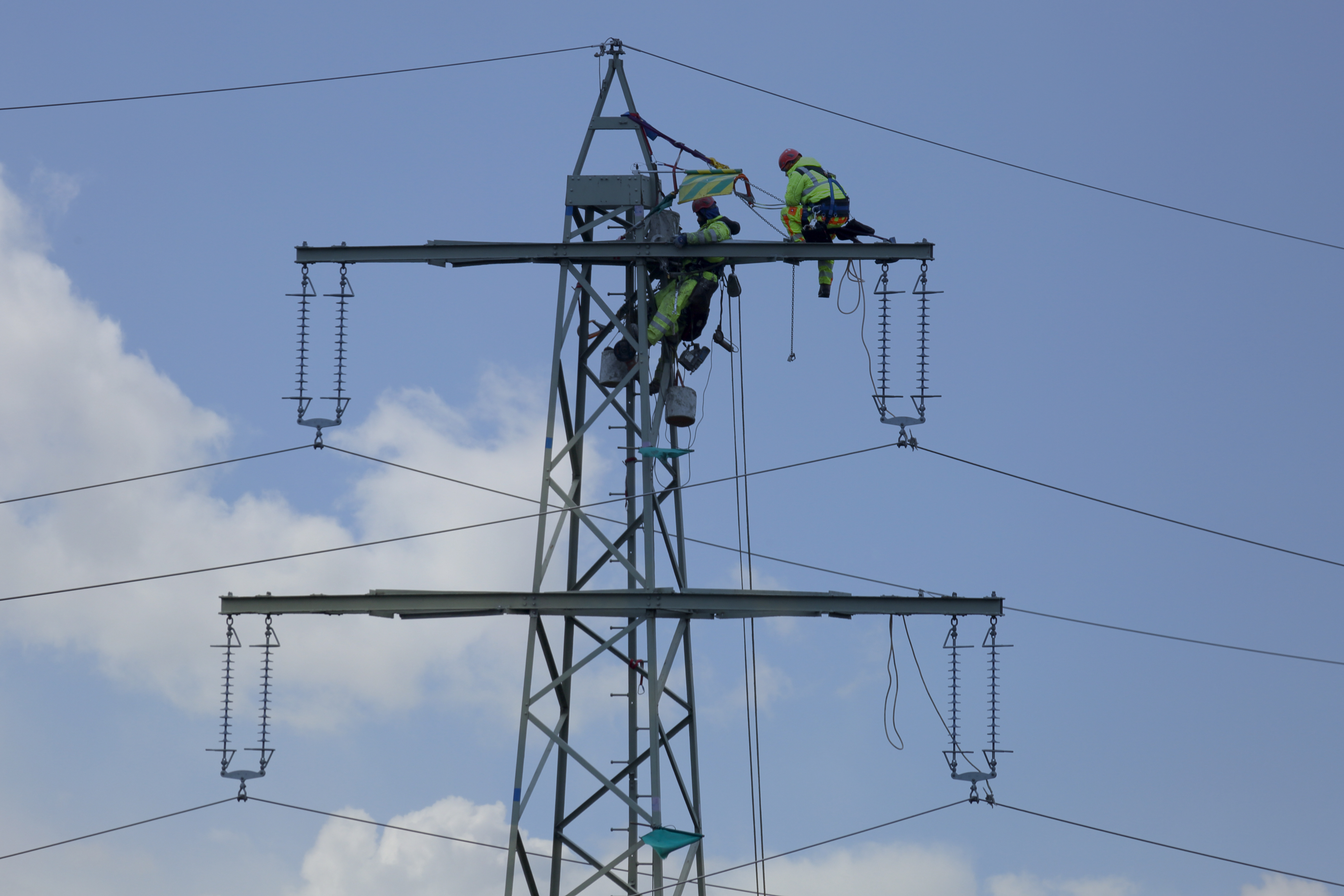 Divi elektroapgādes darbinieki strādā uz augstsprieguma elektropārvades staba zilās debess fonā.