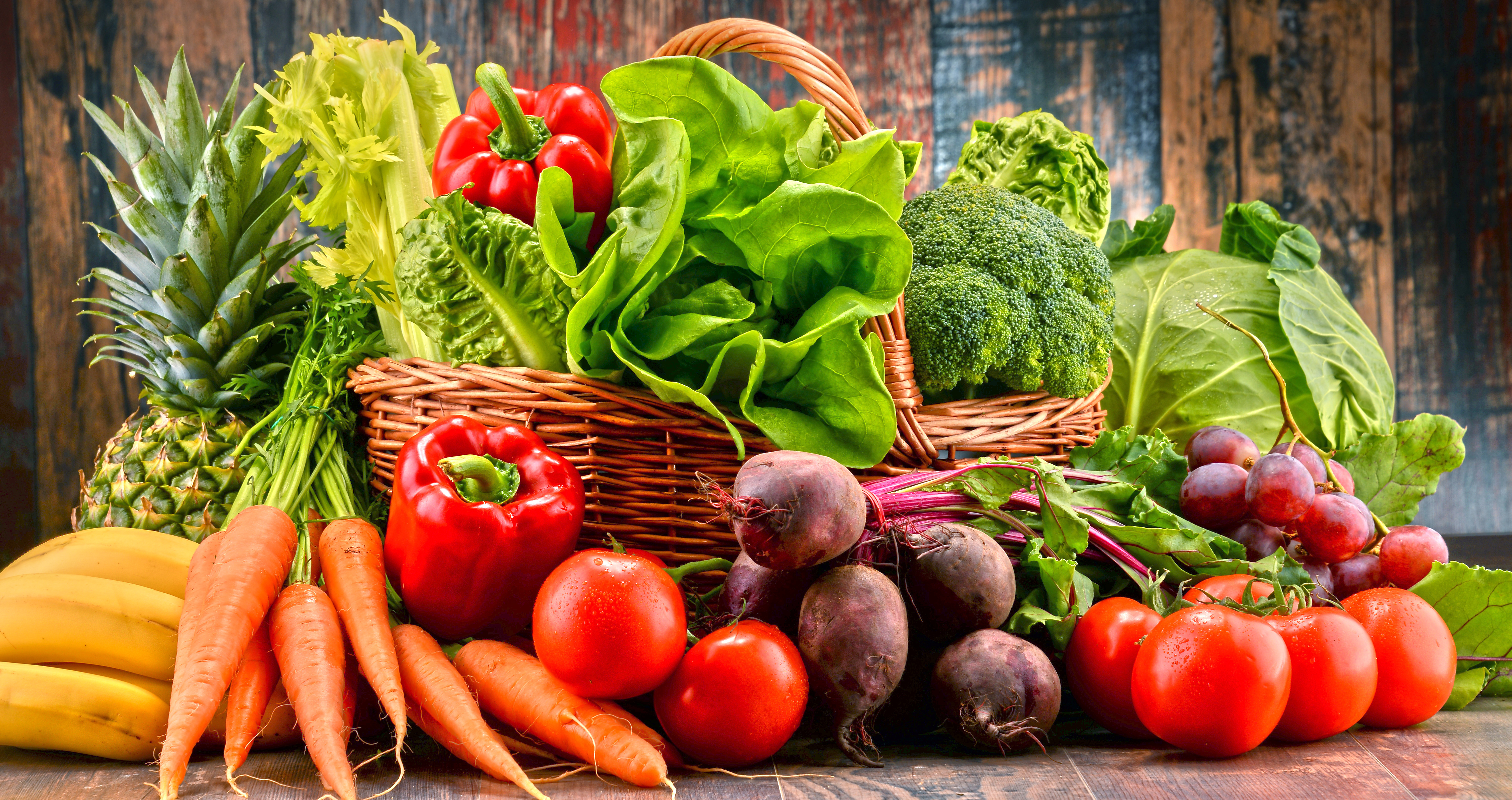 Assorted fresh fruits and vegetables including pineapple, bananas, carrots, tomatoes, bell peppers, lettuce, broccoli, cabbage, beets, and grapes arranged in baskets and on a rustic table.