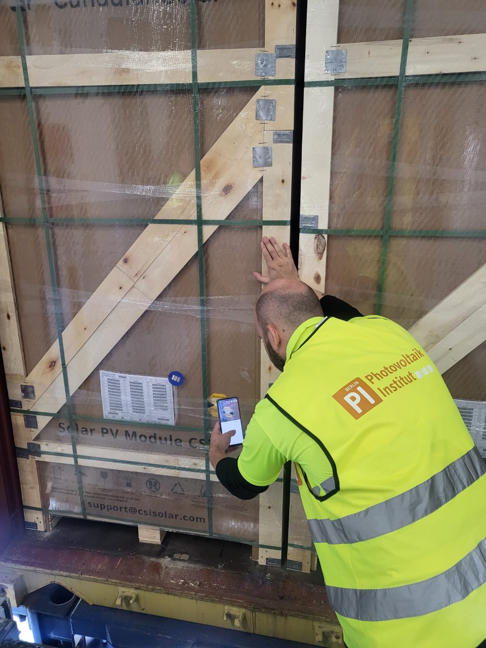 Trabajador inspeccionando un módulo solar embalado en una caja de madera, utilizando un dispositivo móvil, con chaleco de seguridad del Instituto Fotovoltaico.