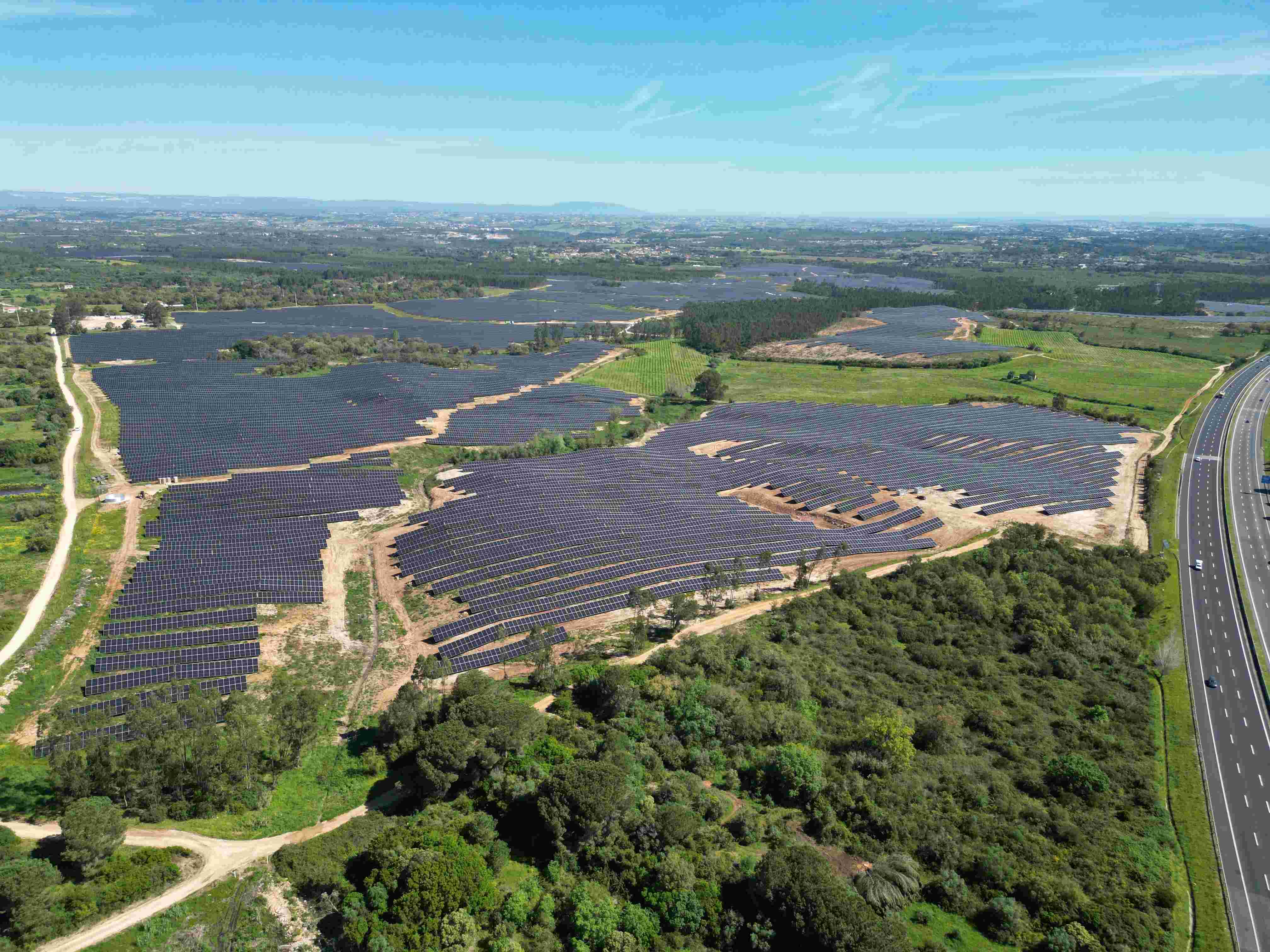 Aerial view of a large solar farm with numerous solar panels spread across the landscape, bordered by green fields and a highway on the right, under a clear blue sky