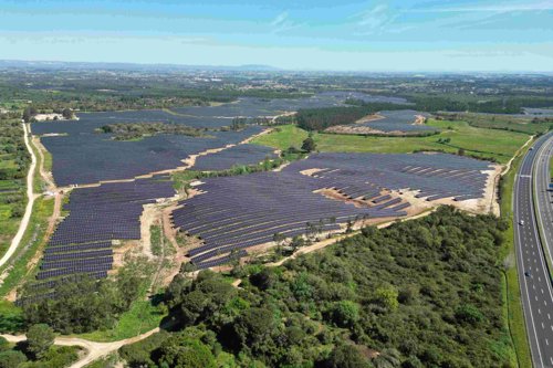 Aerial view of a large solar farm with numerous solar panels spread across the landscape, bordered by green fields and a highway on the right, under a clear blue sky