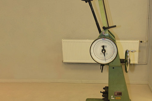 Grön slagprovningsutrustning, med mätare och arm, placerad på ett beige golv framför en ljusgrå vägg.