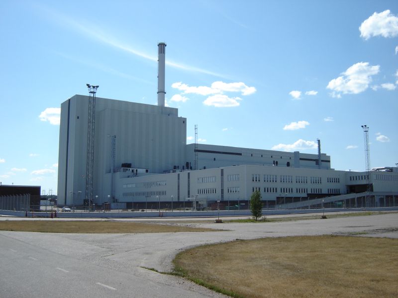 Forsmark kärnkraftverk, reaktor 3, med hög skorsten under blå himmel, sedd från en tom väg och öppen mark.