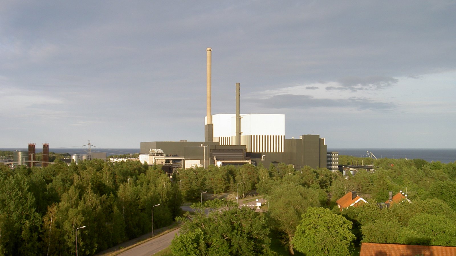 Kärnkraftverket i Oskarshamn med höga skorstenar bakom tät grön skog och några röda hustak vid kusten.
