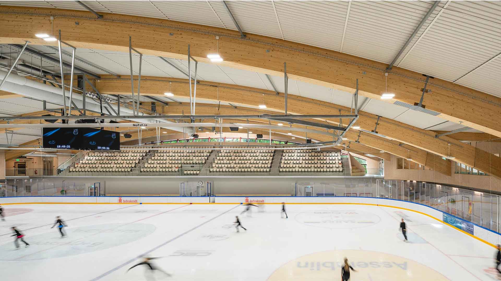 Inomhusbild av en ishall med skridskoåkare på isen, läktare och takbalkar i limträ.
