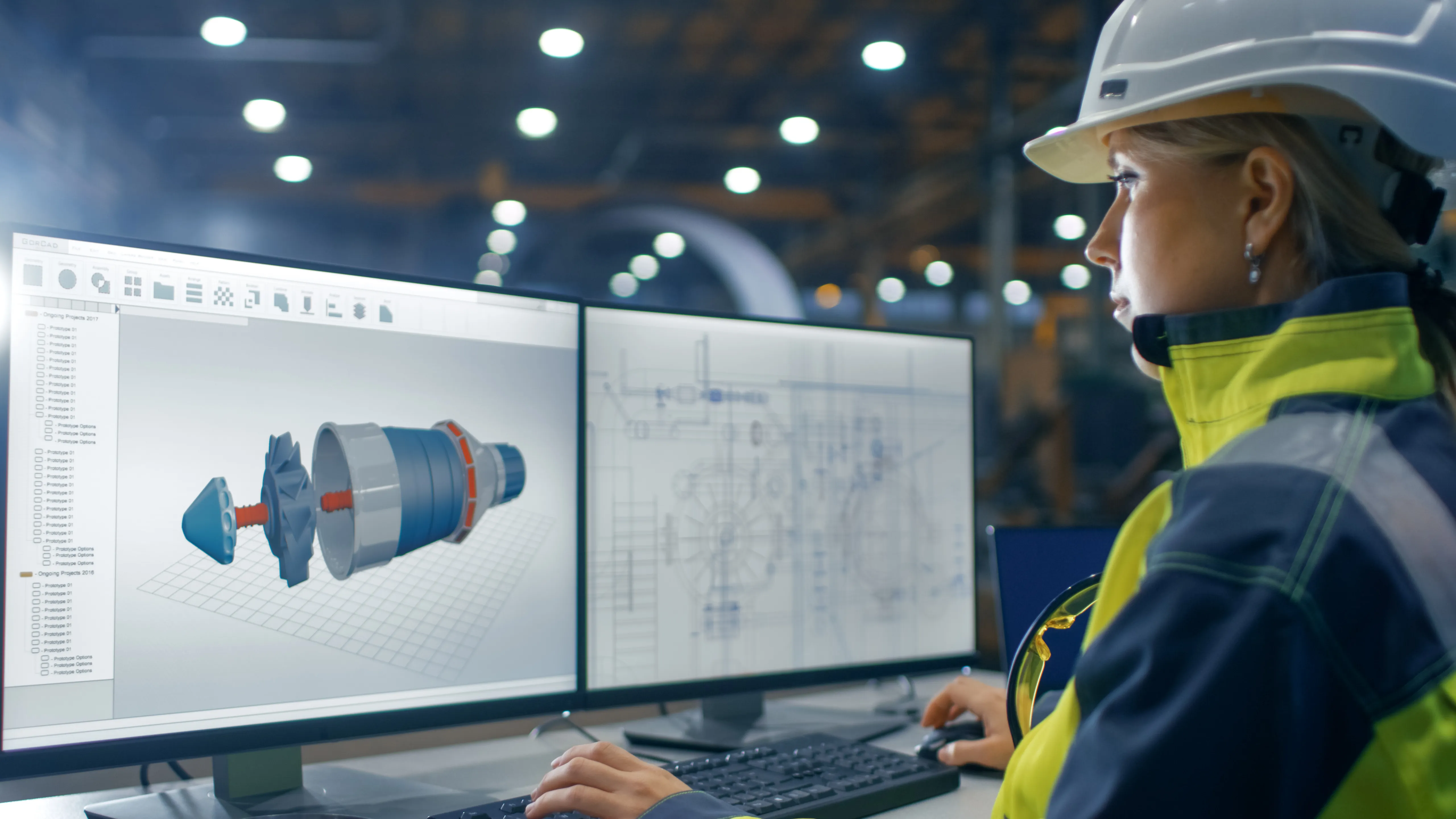 Female Kiwa Engineer in a hard hat reviewing 3D CAD design drawings on dual computer monitors in an industrial workspace